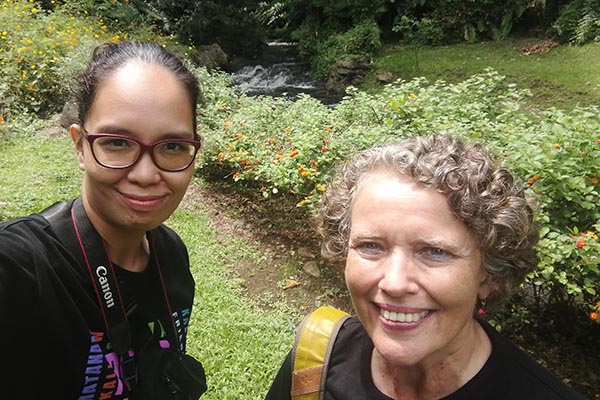 Anna Gingco and Marlies Roth smiling and posing for a selfie in front of a stream and flowering shrubs.